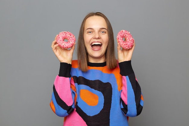 Blanke vrouw die een trui draagt en heerlijke donuts vasthoudt, lacht graag, geeft niet om calorieën, geniet van het eten van een dessert poseren geïsoleerd over een grijze achtergrond