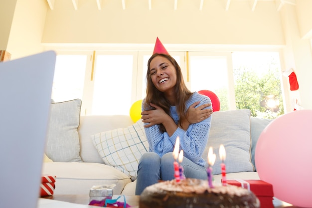 Blanke vrouw die een verjaardagsvideogesprek heeft met een feestmuts. zittend op de bank, verjaardagstaart op de voorgrond. zelfisolatie thuis tijdens de covid 19-coronaviruspandemie.