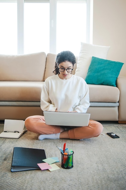 Blanke vrouw die op laptop werkt en met documenten thuis zittend op de vloer quarantaine lifesty...