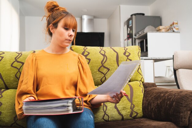 Blanke vrouw die thuis op de bank zit te werken, naar een document kijkt en een map vasthoudt