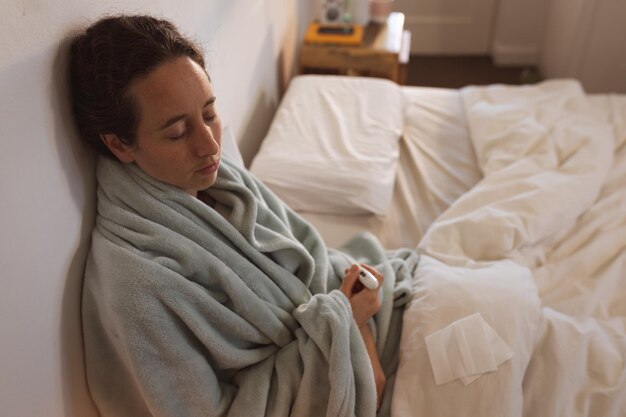 Foto blanke vrouw die tijd thuis doorbrengt, zichzelf isoleert en sociale afstand neemt in quarantaine-afsluiting tijdens de coronavirus covid 19-epidemie, rechtop in bed zit en een thermometer vasthoudt