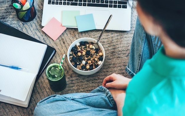 Blanke vrouw doet huiswerk tijdens het eten van granen met vers groen sap op de vloer