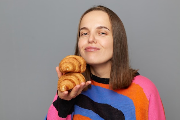Blanke vrouw draagt trui poseren geïsoleerd over grijze achtergrond met heerlijke croissant ruikt van plezier houdt van snoep eten heeft een goede eetlust kijkend naar de camera