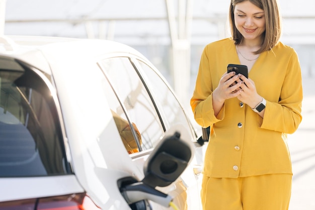 Blanke vrouw gebruikt smartphone tijdens het wachten en stroomvoorziening maakt verbinding met elektrische voertuigen