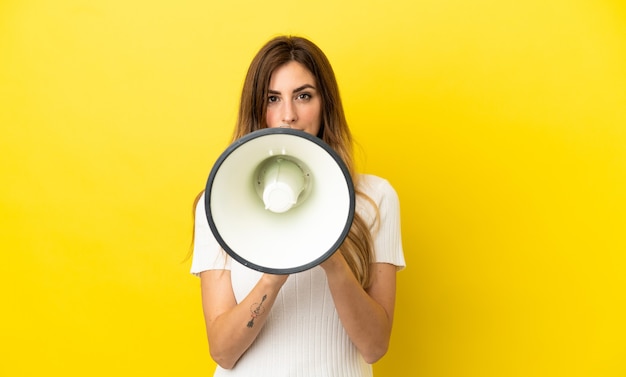 Blanke vrouw geïsoleerd op gele achtergrond die door een megafoon schreeuwt om iets aan te kondigen