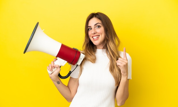 Blanke vrouw geïsoleerd op gele achtergrond met een megafoon en wijzend op een geweldig idee