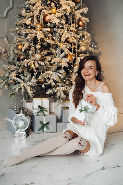 Blanke vrouw in jurk en kousen vormt in de buurt van de kerstboom met veel cadeautjes en glimlachen