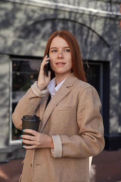 Blanke vrouw in stijlvolle beige jas die belt met smartphone, koffie drinkt