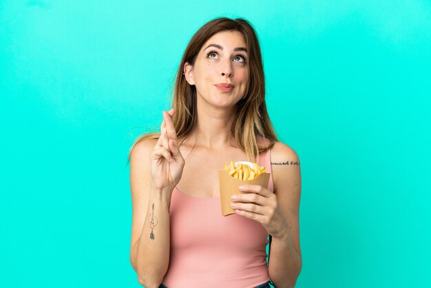 Blanke vrouw met gefrituurde chips geïsoleerd op blauwe achtergrond met vingers die elkaar kruisen en het beste wensen