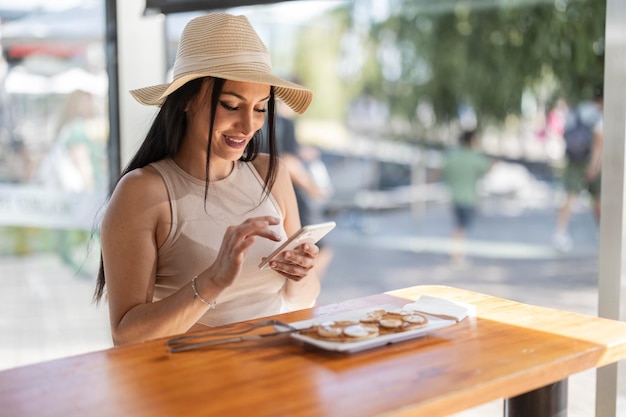 blanke vrouw met hoed in restaurant met ontbijt pannenkoeken maakt foto's van eten met een smartphone