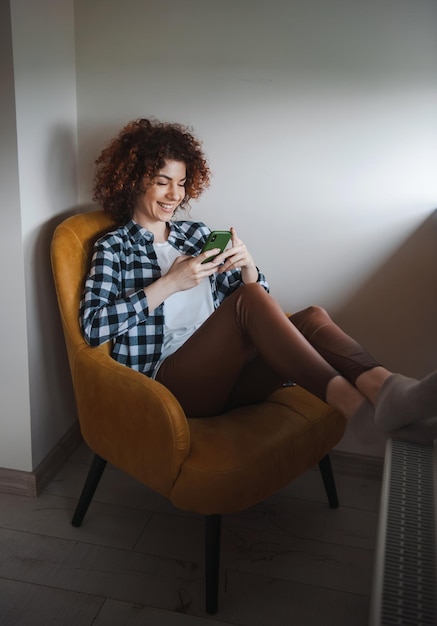 Blanke vrouw met krullend haar zittend in de fauteuil thuis in de ochtend sms-berichten op t...