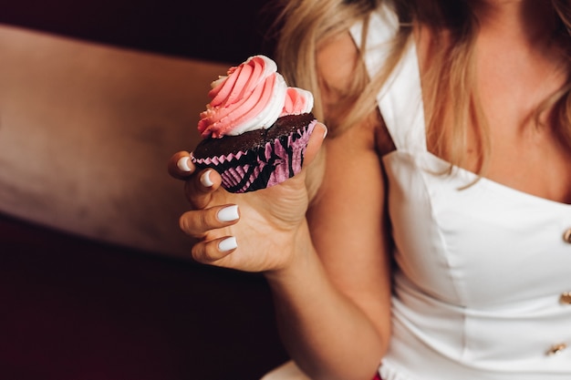 Blanke vrouw met lang blond golvend haar zit op de bank, drinkt koffie en eet lekkere roze cupcakes