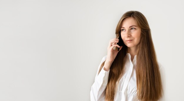 Blanke vrouw met lang bruin haar glimlachend sprekend door smartphone met telefoongesprek op witgrijze achtergrond thuis of op kantoor Bannerkopie ruimte