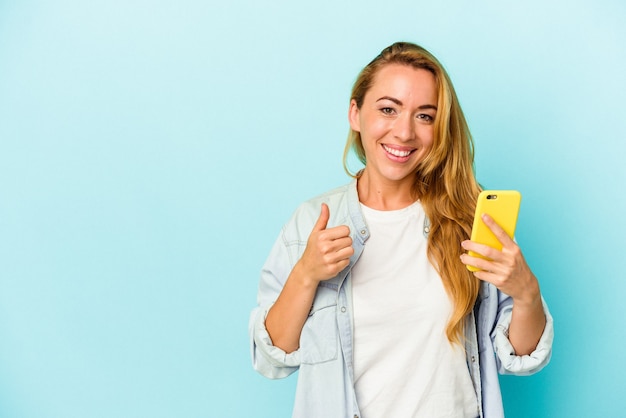 Blanke vrouw met mobiele telefoon geïsoleerd op blauwe achtergrond glimlachend en duim omhoog