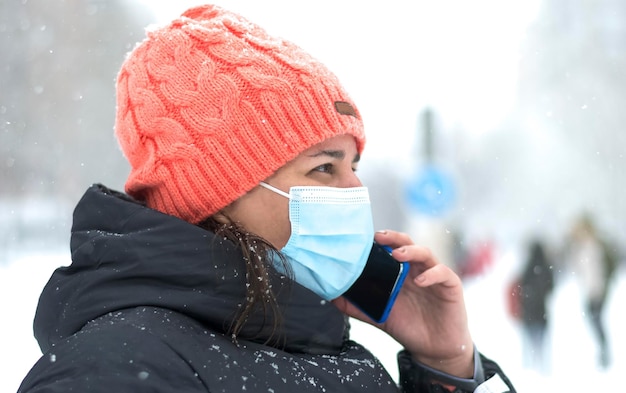 Blanke vrouw met pet en masker voor het coronavirus die tijdens een sneeuwval op haar smartphone praat.