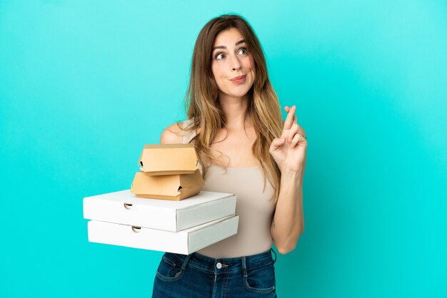 Blanke vrouw met pizza's en hamburger geïsoleerd op een blauwe achtergrond met vingers die elkaar kruisen en het beste wensen