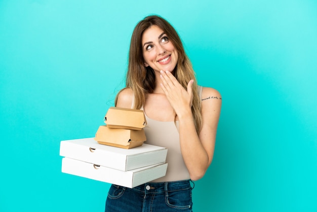 Blanke vrouw met pizza's en hamburger geïsoleerd op een blauwe muur terwijl ze glimlacht