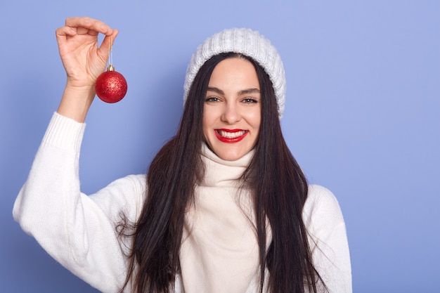Blanke vrouw met rode kerst bal in handen, staat glimlachend geïsoleerd over blauwe muur, brunette vrouw draagt warme witte trui