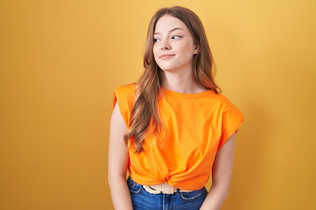 Blanke vrouw staande over gele achtergrond glimlachend naar de zijkant kijkend en weg starend denkend.