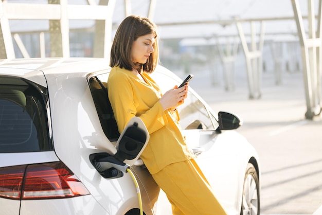 Blanke vrouw staat met mobiele telefoon in de buurt van haar elektrische auto en wacht wanneer het voertuig wordt opgeladen