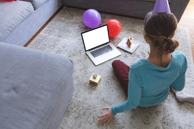 Foto blanke vrouw thuis in de woonkamer viert haar verjaardag met ballonnen, draagt een feestmuts en voert een videogesprek op laptop. sociale afstand tijdens de quarantaine-afsluiting van covid 19.