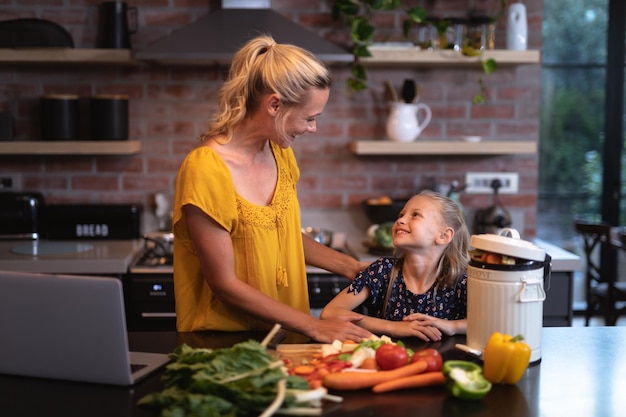Blanke vrouw thuis, met haar dochter aan een tafel staan, laptop gebruiken, groenten snijden, sociale afstand nemen en zelfisolatie in quarantaine tijdens de covid19-epidemie van het coronavirus