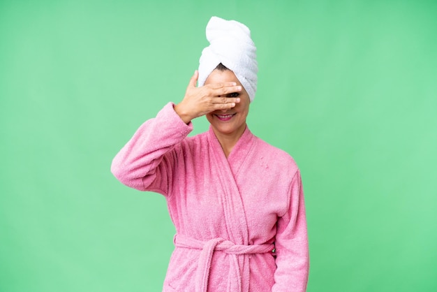 Blanke vrouw van middelbare leeftijd in een badjas over een geïsoleerde achtergrond die de ogen met de handen bedekt en glimlacht