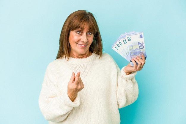 Blanke vrouw van middelbare leeftijd met bankbiljetten geïsoleerd op een blauwe achtergrond en met de vinger naar je wijzend alsof uitnodigend dichterbij komt.