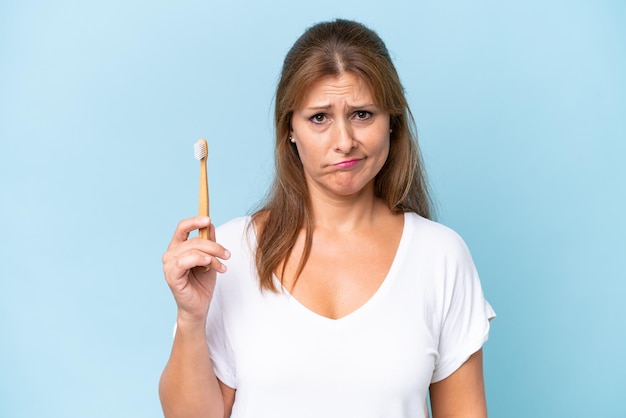 Blanke vrouw van middelbare leeftijd tanden poetsen geïsoleerd op blauwe achtergrond met droevige uitdrukking