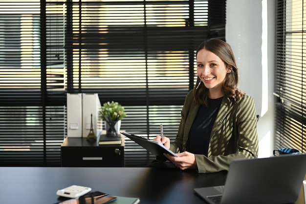 Blanke vrouwelijke corporate CEO in slimme kleding met klembord werkend met laptopcomputer in haar kantoor