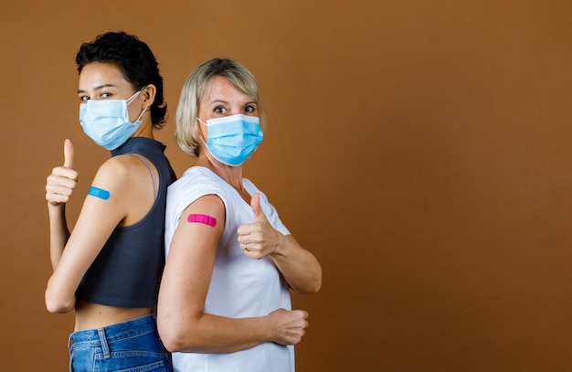 Blanke vrouwelijke patiënten dragen een gezichtsmasker terwijl ze naar de camera kijken, op elkaar leunen en de duim samen met kleurrijke pleister tonen na vaccinatie voor een bruine muurachtergrond.