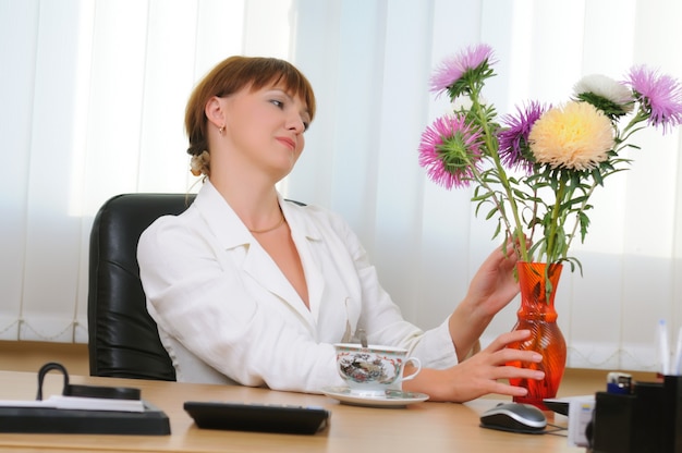 Foto blanke witte brunette vrouw zit aan het bureau bloemen boeket knuffelen. beker, computermuis, map met documenten op tafel.