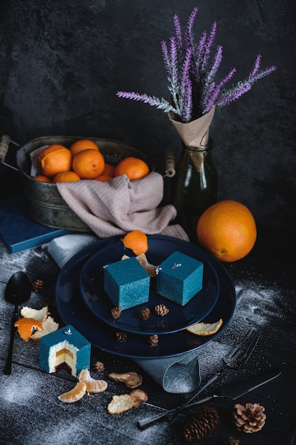 Blauwe cake gevuld met sinaasappel en mandarijn in de vorm van een kubus. Vlakbij op een zwarte tafel staan ingrediënten sinaasappels, mandarijnen, noten. Rustieke stijl