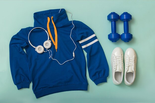 Blauwe hoodie met koptelefoon, halters en witte sneakers op een blauwe achtergrond. sportieve lifestyle accessoires.