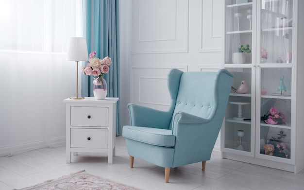 Blauwe klassieke fauteuil en witte ladekast met tafellamp en bloemenvaas in witte kamer