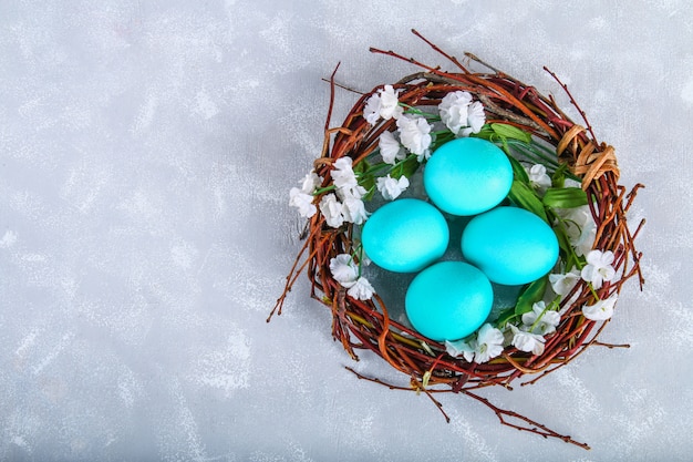 Blauwe paaseieren in een nest met witte bloemen op een grijze concrete achtergrond.