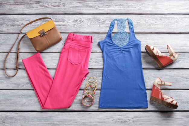 Blauwe top en roze broek. Tweekleurige portemonnee en sandalen met sleehak. Trendy zomerkleding voor dames. Bijouterie en kleding op plank.