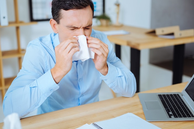 Bless you. Frustrated man sitting at his workplace and putting elbows on the table while keeping eyes closed