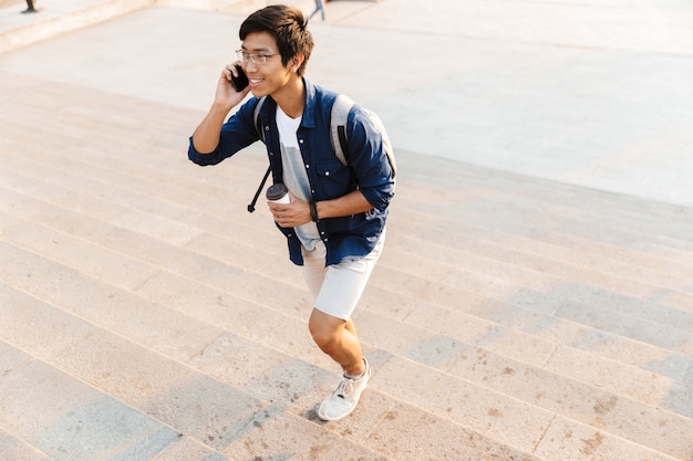 Blij Aziatische mannelijke student in brillen praten door smartphone tijdens het lopen op trappen buitenshuis