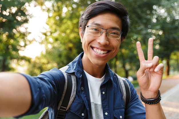 Blij Aziatische mannelijke student in brillen selfie maken en vredesgebaar tonen tijdens het kijken naar de camera buitenshuis