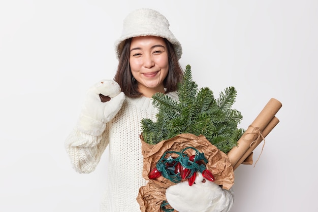 Blij aziatische vrouw draagt winter panama gebreide trui en wanten draagt nieuwjaar attributen voor decoratie heeft feestelijke stemming poses tegen een witte achtergrond. gelukkig wintervakantie concept.