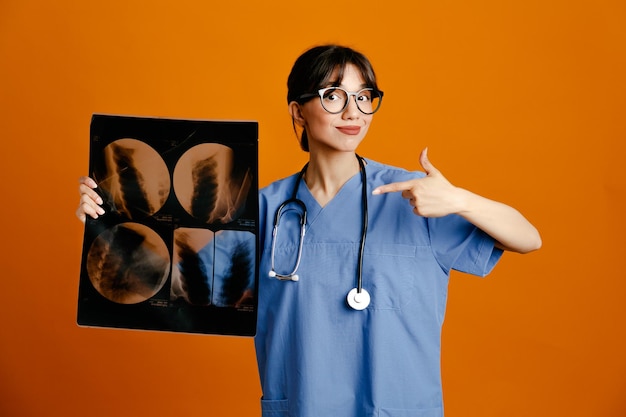 Blij bedrijf en wijst naar xray jonge vrouwelijke arts met uniforme stethoscoop geïsoleerd op een oranje achtergrond