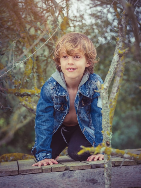 Foto blij blond kind in spijkerjasje glimlachend en wegkijkend terwijl hij op een houten pad kruipt te midden van bemoste boomtakken in het bos