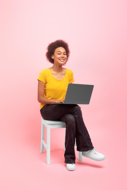 Blij dat de jonge afro-amerikaanse curly lady blogger op een stoel zit te typen op een laptop geïsoleerd op een roze achtergrond