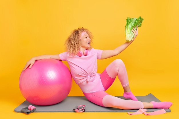 Blij dat gezonde sportieve vrouw groene groente vasthoudt, houdt zich aan het dieet leidt actieve levensstijl heeft regelmatige training leunt op fitball geïsoleerd op gele achtergrond Dieet welzijn en sport concept