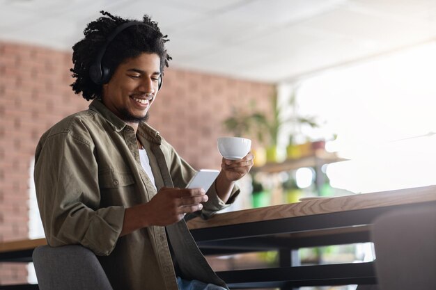 Blij dat jonge Afro-Amerikaanse krullende man in draadloze koptelefoon naar smartphone aan tafel kijkt