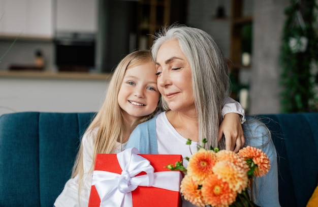Blij dat kaukasische kleindochter knuffelt senior vrouw geeft bloemendoos met cadeau feliciteert veel plezier