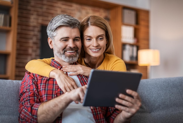 Blij dat volwassen europese dame die man met tablet knuffelt, geniet van winkelen op afstand zittend op de bank in het interieur van de woonkamer