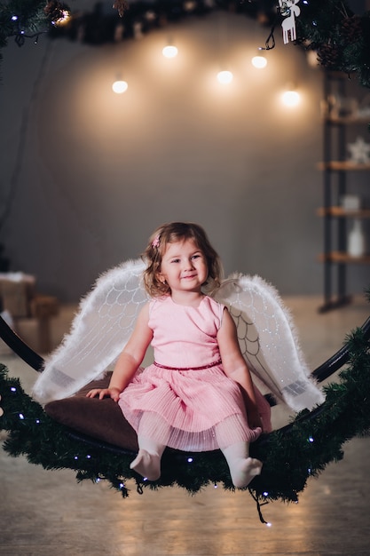 Blij en gelukkig kind zittend en poseren op dennenkrans versierd met lichte garland. Aanbiddelijk meisje dat in leuke roze kleding draagt en erachter engelenvleugels heeft. Kid glimlachend en camera kijken.