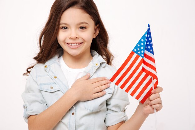 Blij glimlachend meisje dat de Amerikaanse vlag vasthoudt en haar borst aanraakt terwijl ze liefde uitdrukt en staat geïsoleerd in een witte muur